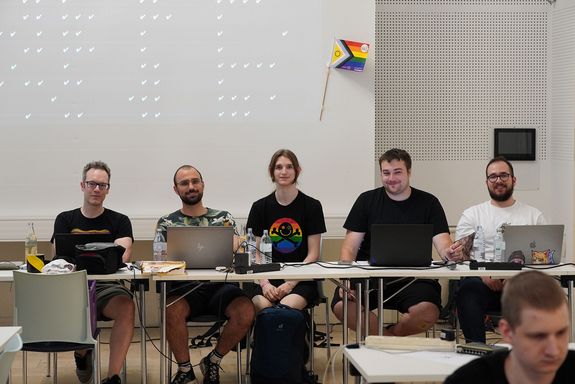 Fünf Personen sitzend bei Tisch mit Blick zur Kamera