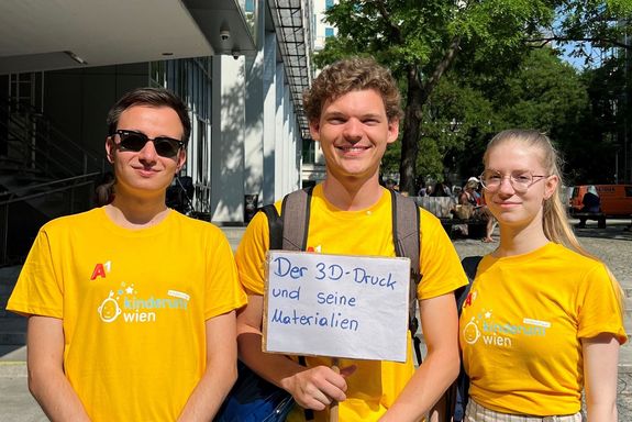 Drei Studierende mit einem orangen, gebrandeten Kinderuni T-Shirt halten ein Schild mit der Aufschrift der 3D Druck und seine Materialien