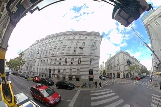 Eine Straßenszene in der Stadt mit Zebrastreifen, einem roten Auto und Gebäuden. In der grauen Box am Dach des Autos befinden sich die Millimeterantennen. Fotografiert mit Fischaugen-Objektiv.