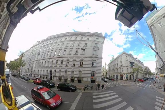 Eine Straßenszene in der Stadt mit Zebrastreifen, einem roten Auto und Gebäuden. In der grauen Box am Dach des Autos befinden sich die Millimeterantennen. Fotografiert mit Fischaugen-Objektiv.
