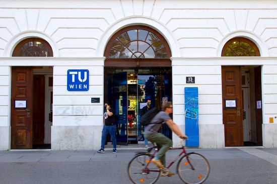 Entrance area of ​​the TU Wien Campus Karlsplatz. Two people come out wearing masks, a cyclist drives past.