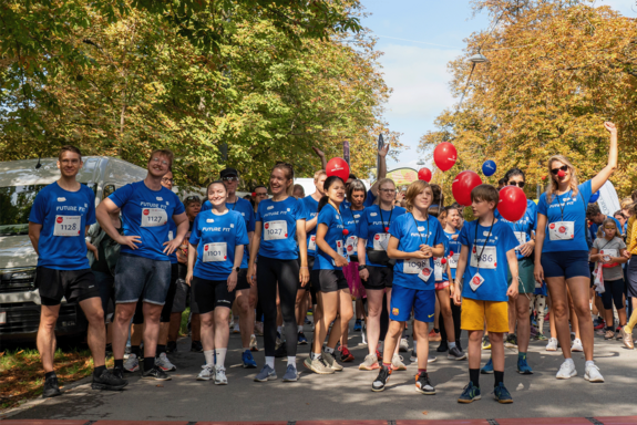 Läufer_innen vor dem Start mit roten Luftballons, manche mit Roten Nasen. Kinder und Erwachsene, alle tragen blaue TUW-T-Shirts.