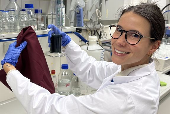 a woman in a white coat wearing safety goggles, holding a piece of cloth in a chemistry lab