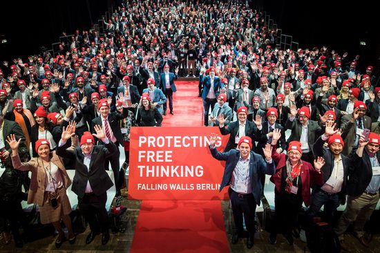 Der Blick von der Bühne in ein fröhliches Publikum, alle tragen rote Mützen und heben stehend ihre Arme. In der Mitte ein roter Gang mit dem Slogan "Protecting Free Thinking".