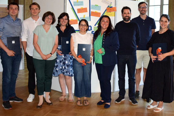 Gruppenfoto mit neun Personen, die meisten von ihnen halten eine Urkunde in Hönden. Sie tragen sommerliche Kleidung