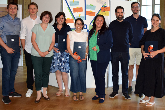Gruppenfoto mit neun Personen, die meisten von ihnen halten eine Urkunde in Hönden. Sie tragen sommerliche Kleidung