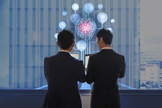 Zwei Geschäftsmänner stehen vor Laptops und betrachten Daten auf ihren Bildschirmen, während im Hintergrund ein abstraktes digitales Netzwerk mit einer zentralen Gehirn-ähnlichen Struktur dargestellt wird, das moderne Technologien und Datenanalyse symbolisiert.