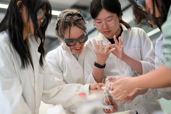 Mädchen mit Schutzbrillen und weißen Labormänteln bei einem Experiment.