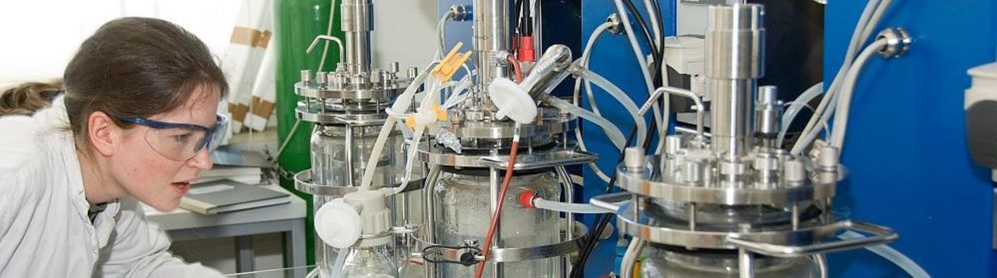 Eine Wissenschaftlern mit Schutzbrille steht vor drei Glaskolben, an denen viele Schläuche hängen