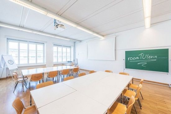 Klassenzimmer mit weißen Tischen und braunen Sesseln, rechts vorne eine grüne Tafel mit dem Schriftzug roomTUlearn in Kreide geschrieben