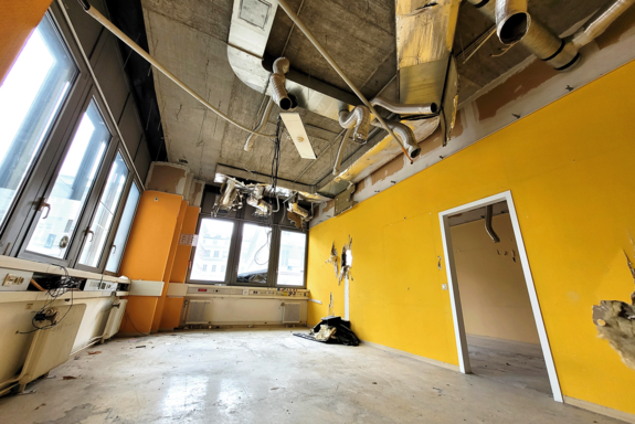 Raum im Weitwinkel aufgenommen. Rechts gelbe Wand, links Fensterfront mit orangen Pfeilern. Die Decke ist abgehängt, u.a. Lüftungsrohre sichtbar.
