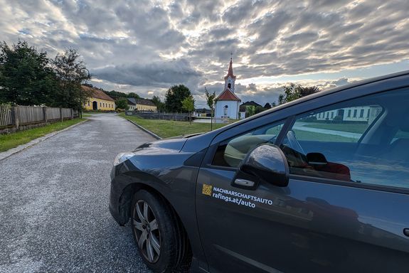 Auto mit Aufschrift "Nachbarschaftsauto, rafings.at/auto". Danaben eine Straße und im Hintergrund Häuser und eine Kirche
