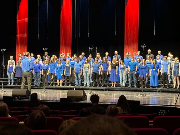 Der TU Wien Chor während des Wettbewerbsauftritts bei den European Choir Games 2025: Sänger_innen sind blau-schwarz gekleidet und stehen in gemischter Aufstellung auf der Bühne. Dahinter sieht man Rote Banner von der Decke hängen. Auf der Bühne stehen Mikrofone. Vor dem Chor steht der Chorleiter mit dem Rücken zum Publikum, das man im Vordergrund des Bildes erkennen kann.