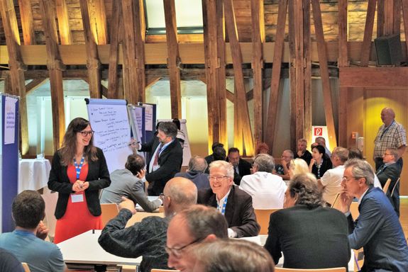 Gruppe an menschen sitzend an Tischen, mehrere Personen stehend, Flipcharts