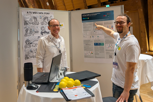 The photo shows two people at a small exhibition stand with poster walls and a high table with laptops, flyers, and advertising material.