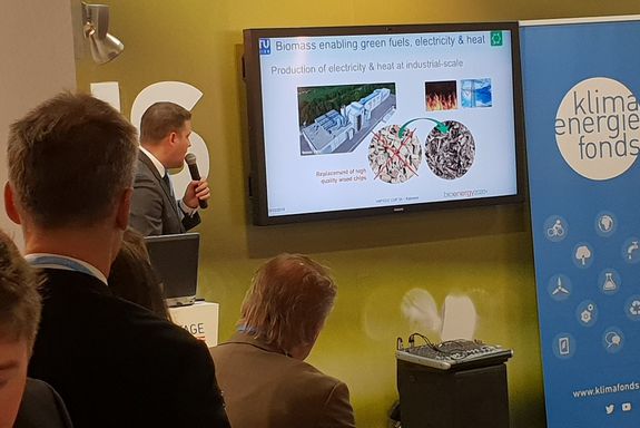 Dr. Stefan Müller (TU Wien) infront of Flatscreen presenting "Biomass enabling green fuels, electricity and heat", some people watching, listening to himlistening
