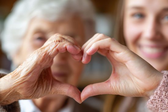 Herz aus Händen groß im Vordergrund, im Hintergrund unscharf Personen, links ältere Frau, rechts junge Dame
