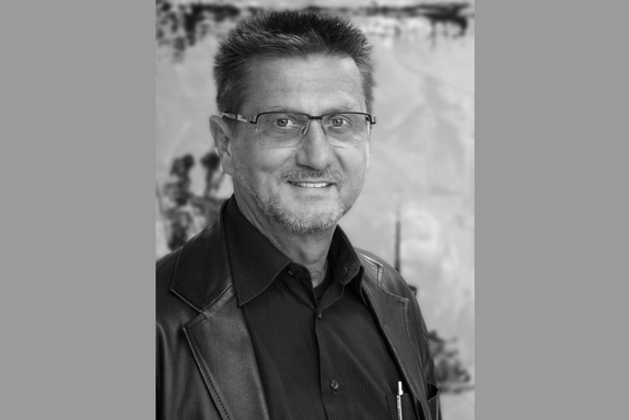 Porträtbild in Schwarz-Weiß. Die Person hat eine Kurzhaarfrisur, trägt Brille, ein schwarzes Hemd und Lederjacke. Hochformat und graue Balken links und rechts.