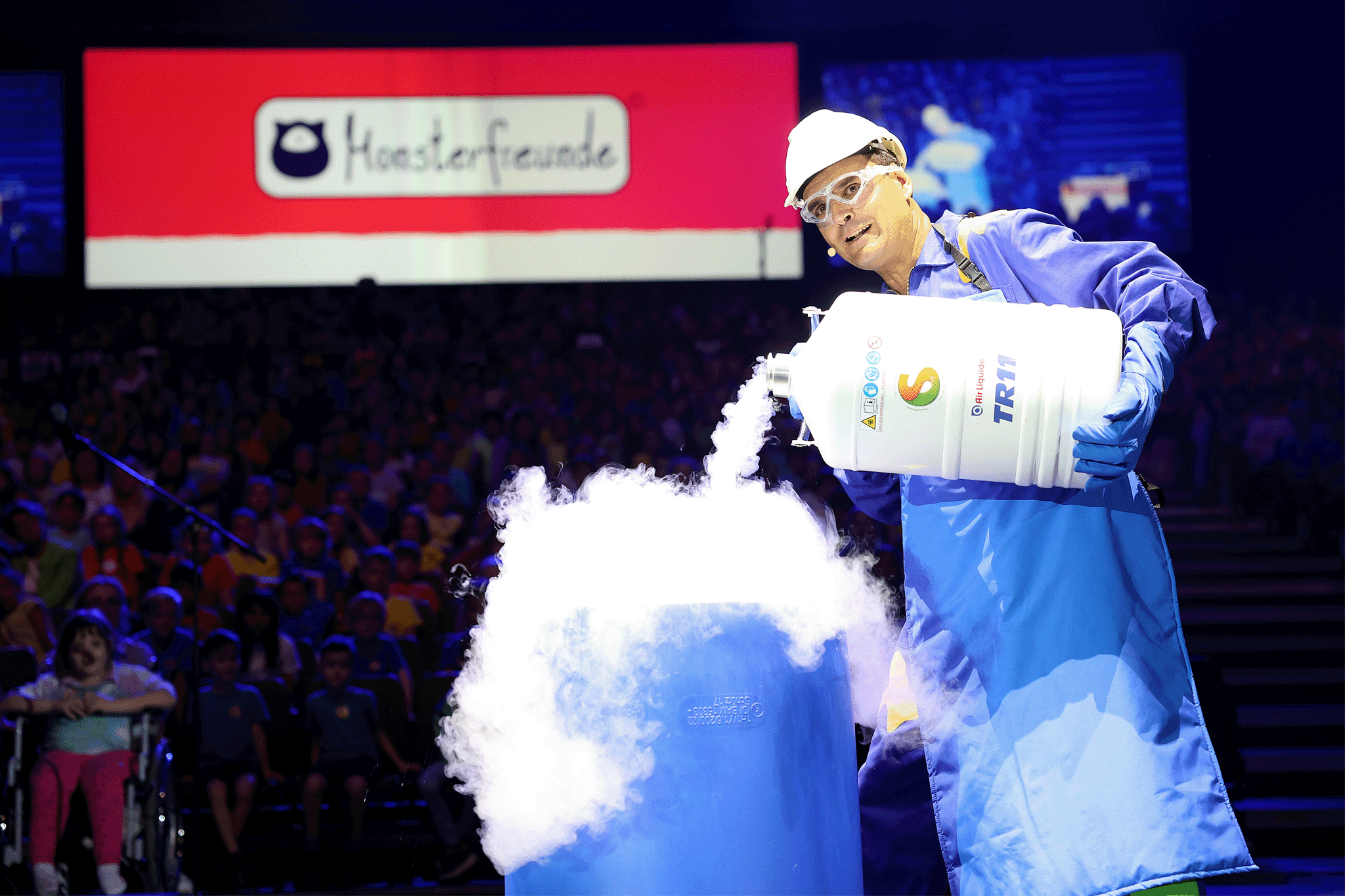 Bernhard Weingartner mit blauem Mantel schüttet eine rauchende Substanz in einen großen blauen Behälter. Die Substanz wabert nach oben. Links oben "Monsterfreunde"-Schriftzug auf Rot.