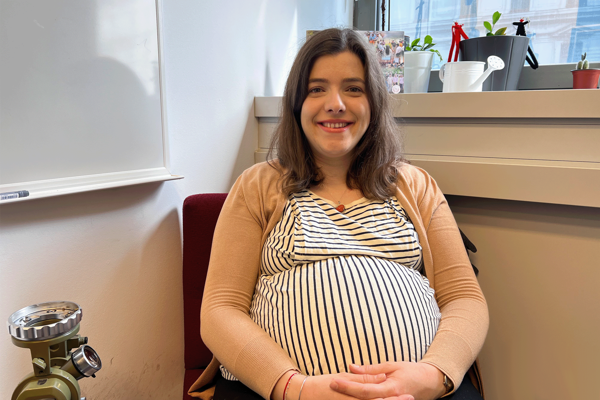Sitzende lächelnde Frau, mit gestreiftem Oberteil und beiger Weste. Daneben eine weiße Tafel. Sie ist schwanger