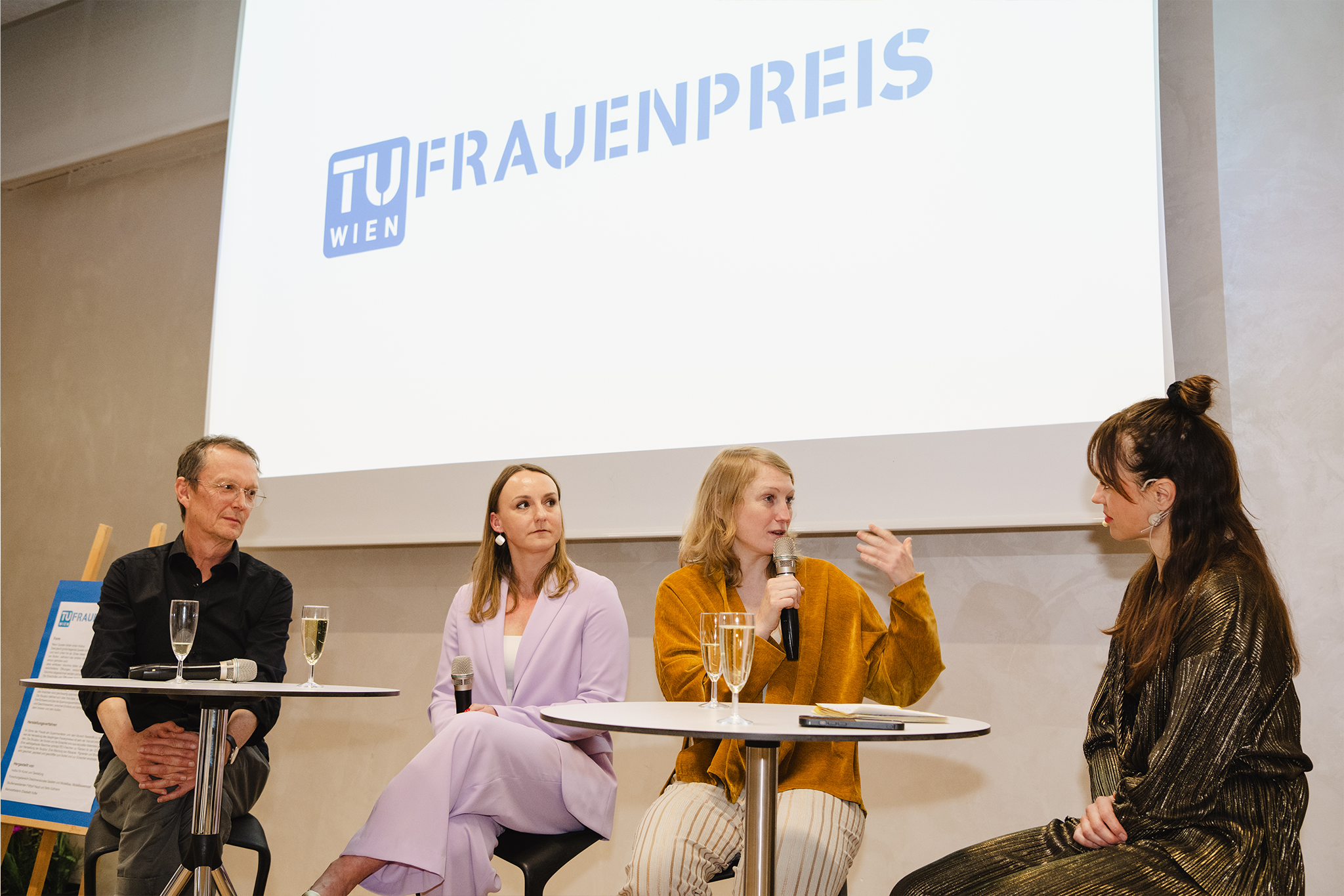Im Hintergrund ein Screen mit dem TUW-Logo und der Schrift "FRAUENPREIS", an zwei Stehtischen auf hohem Hocker vier Personen, die miteinander diskutieren