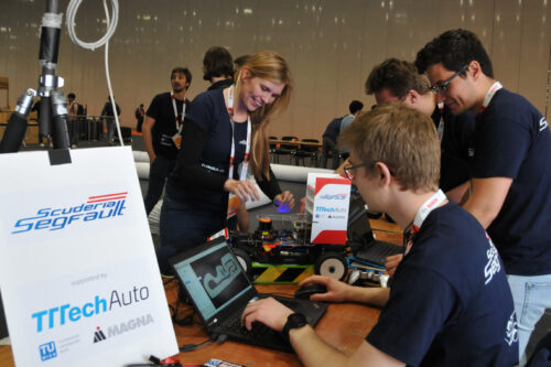 Students working on notebook, with another student and a model car in the background