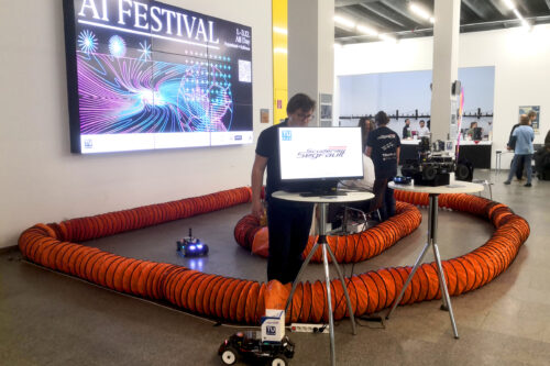 Screen, racing cars on the track and sign of AI festival in the background.