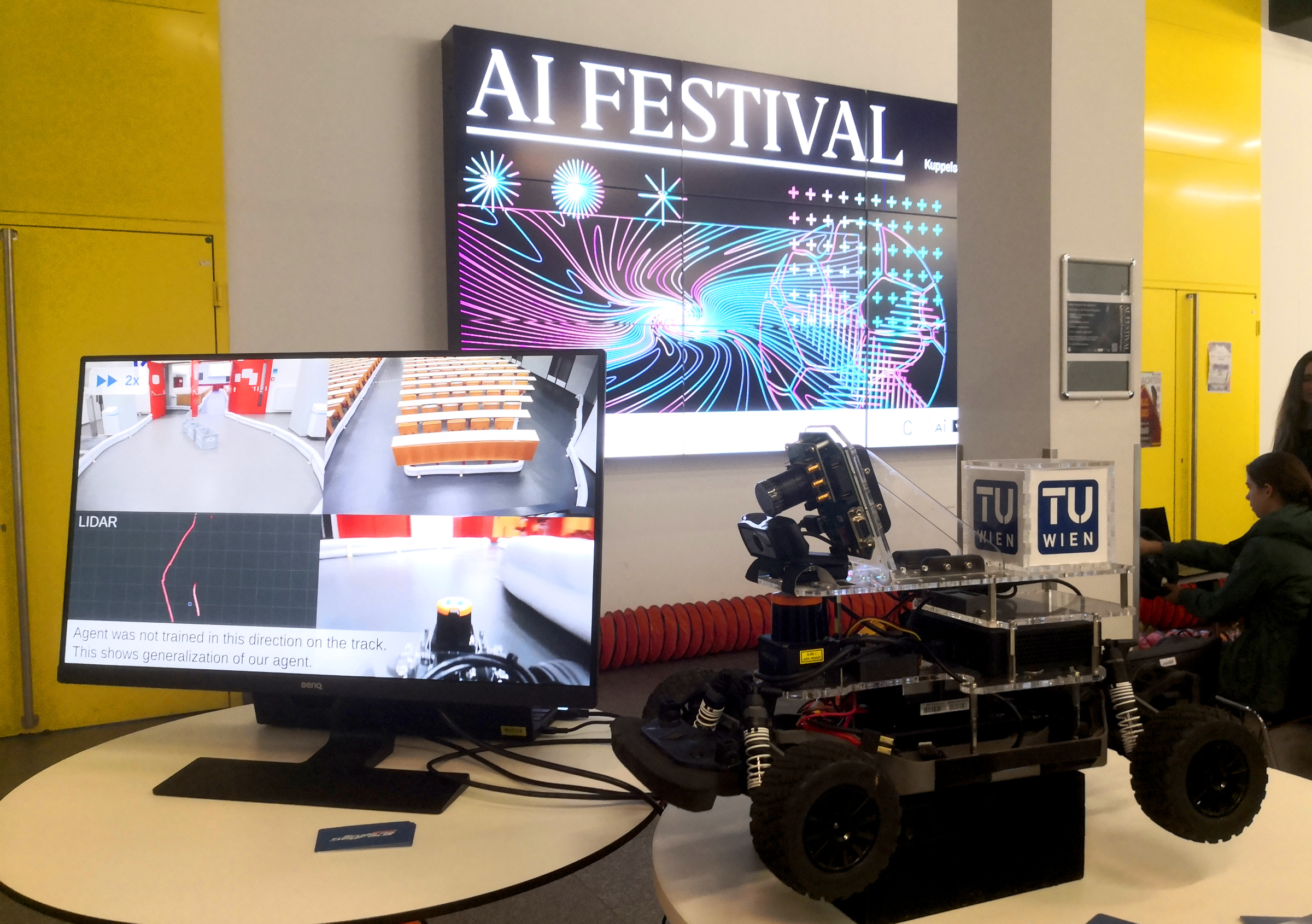 Screen, racing car and sign of AI festival in the background.