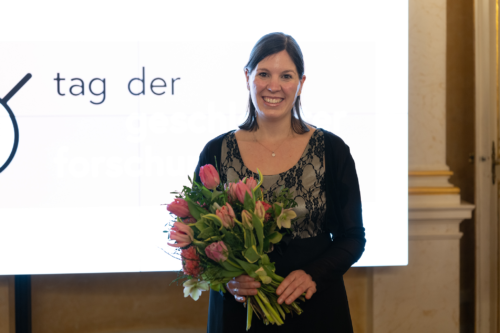 Ruth Strobel lachend mit Blumenstrauß in der Hand, im Hintergrund weißes Leinwand