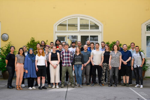 Gruppenfoto Personen stehend vor gelben Haus, großes Fenster in der Mitte, Sträucher im Hinetrgrund sichtbar