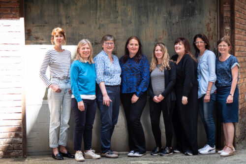 Teamfoto, 8 Personen stehend, außen vor grauer Wand